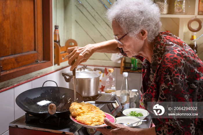 亚洲老年女性在家里的厨房里煮煎蛋，亚洲老年女性的生活方式