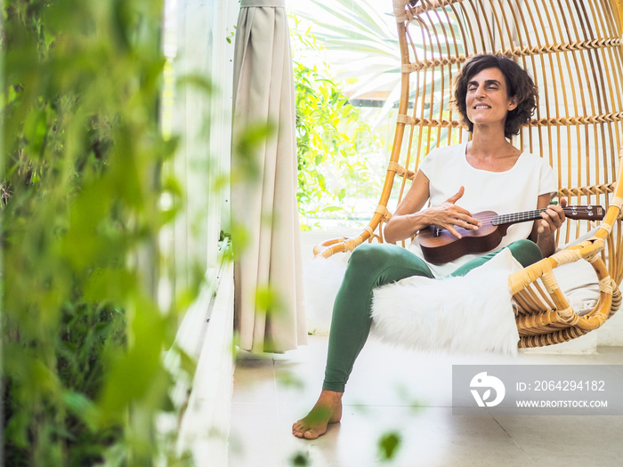 happy and relaxed woman playing ukulele sitting in a bamboo rattan hanging swing chair with green pl