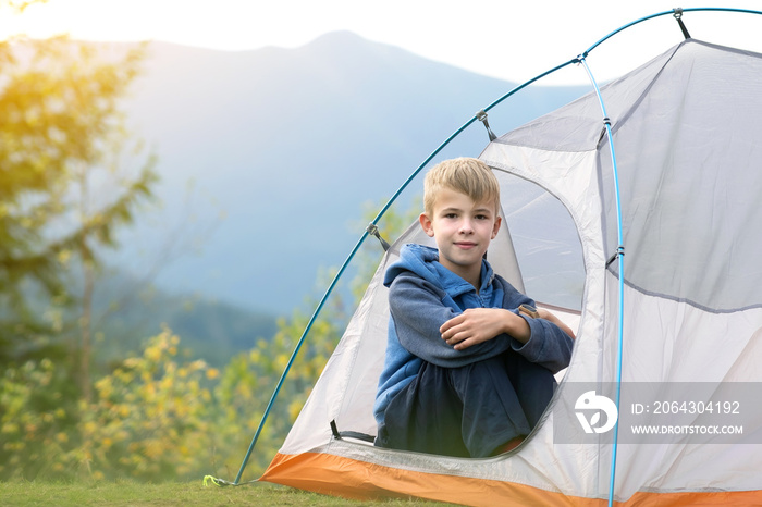 Happy child boy resting in a tourist tent at mountain campsite enjoying view of beautiful summer nat