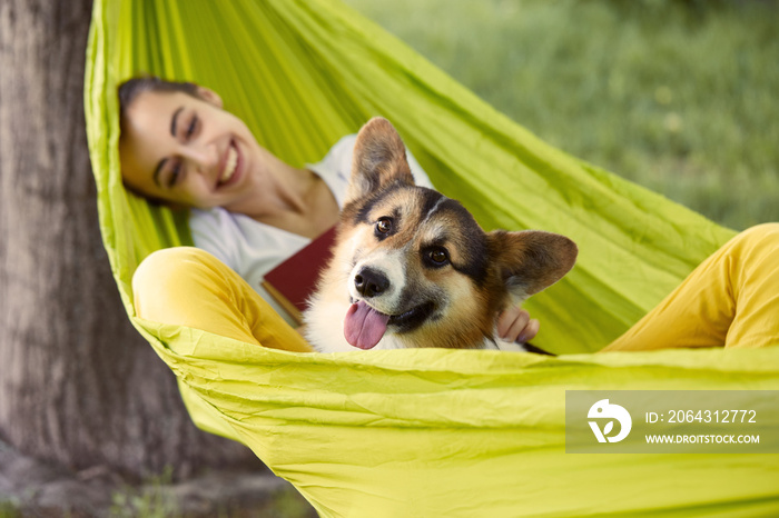 Smiling young woman resting in green hammock with cute dog Welsh Corgi in a park outdoors. Beautiful