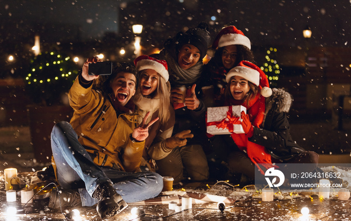 一群快乐的年轻朋友晚上戴着圣诞帽坐在户外喝咖啡。