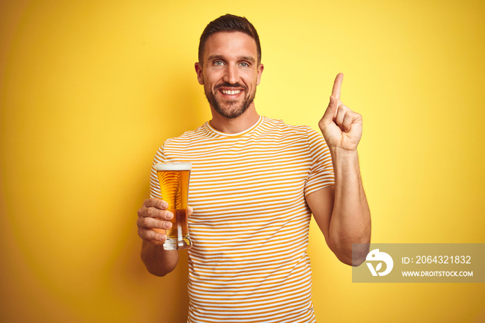 Young handsome man drinking a pint glass of beer over isolated yellow background very happy pointing