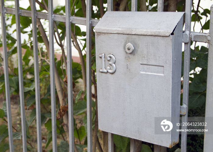 One gray metal mailbox hanging on a fence. The number  13  is stamped on the postbox.