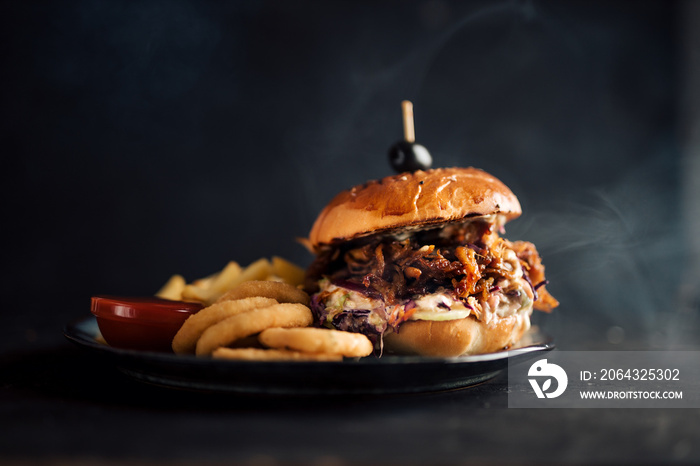 Juicy pulled pork burger on a plate with fries and onion rings. Dark background, copy space.