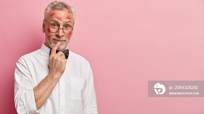 Curious pensioner has thick grey beard keeps finger on lips, looks with interest, dressed in formal 