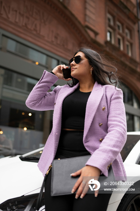 Young businesswoman leaning against luxury car and using phone