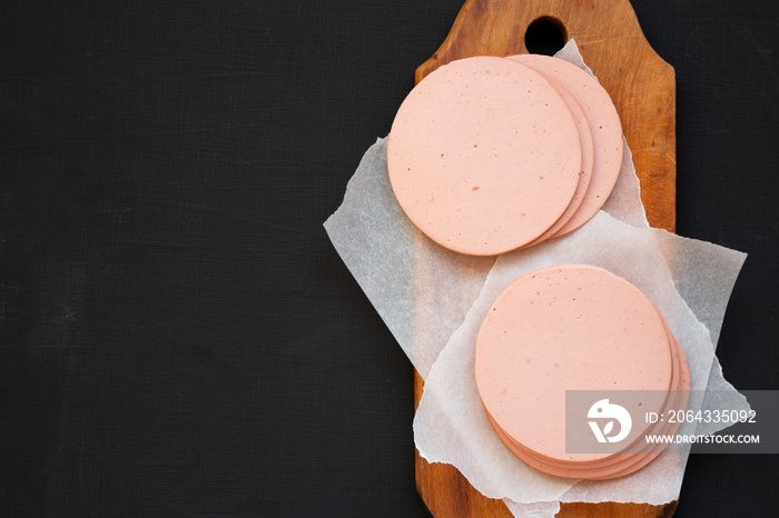 Sliced pork bolgna meat on a rustic wooden board on a black surface, top view. Flat lay, overhead, f