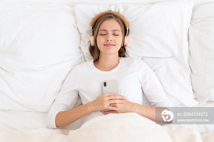 Young female dressed in white spending late morning in bed, listening to music or audiobook from her