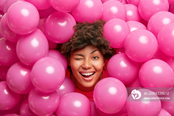 乐观的生日女孩玩得很开心，眨眼微笑，对着许多粉红色的气球摆姿势