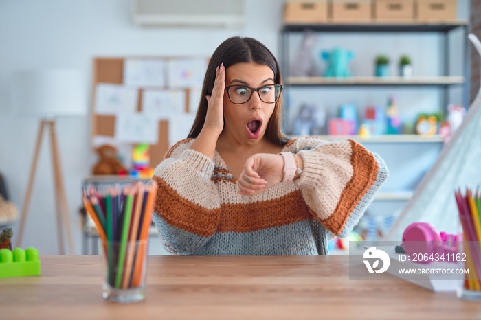 Young beautiful teacher woman wearing sweater and glasses sitting on desk at kindergarten Looking at