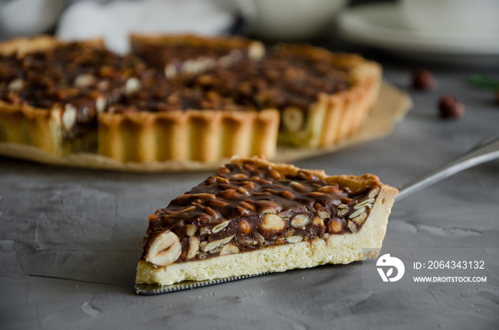 Tart with chocolate caramel, hazelnuts, peanuts, almonds and seed mix on a dark concrete background.