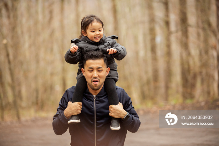 Father carrying happy daughter on shoulders while standing on road against bare trees in forest