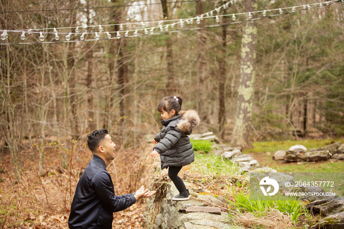 Side view of daughter jumping towards happy father while standing on retaining wall in forest