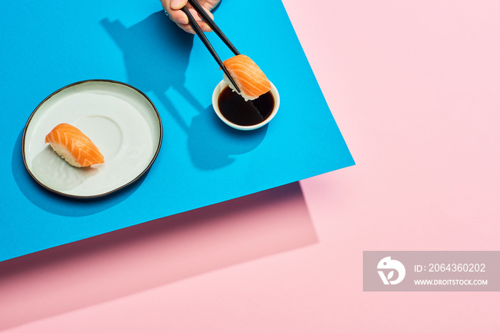 cropped view of woman putting fresh nigiri with salmon into soy sauce on blue, pink background