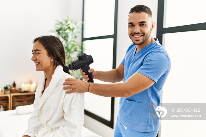 Latin man and woman wearing physiotherapy uniform massaging neck using percussion gun at beauty cent