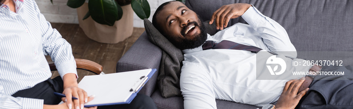 horizontal image of joyful african american man talking while lying on sofa near psychologist holdin