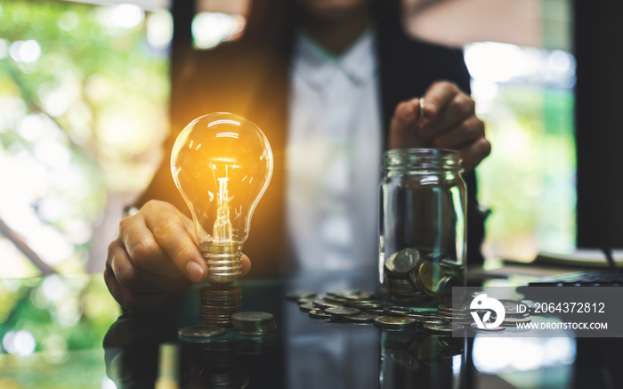 Businesswoman holding a light bulb over coins stack on the table while putting coin into a glass jar