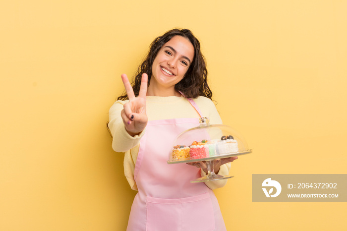 young pretty hispanic woman smiling and looking friendly, showing number two. chef and cakes concept