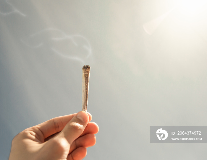 Man holding a lit joint of marijuana with a soft sky and shiny sun in the background. Cigarette with