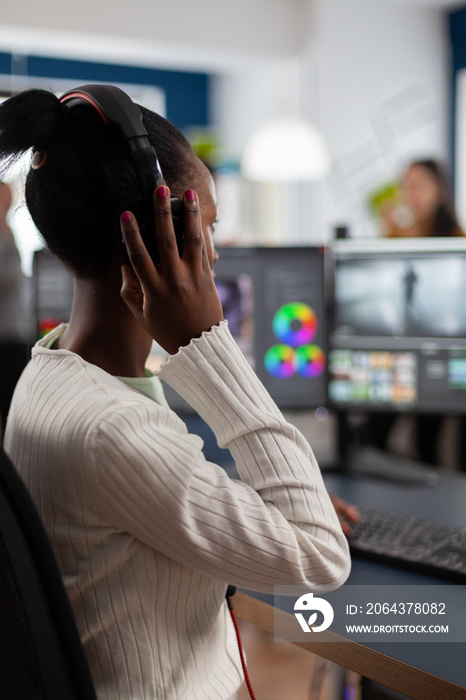 Black video editor working with footage on pc with two monitors, putting on headphones. African vide