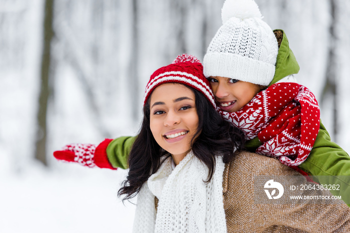 attractive african american mother piggybacking cheerful daughter, smiling and looking at camera in 