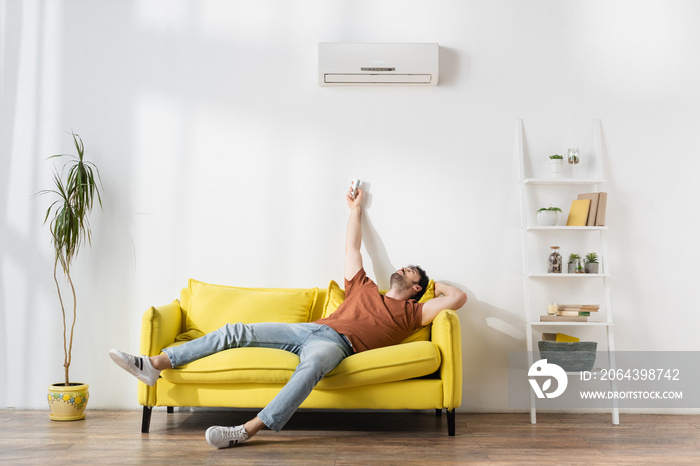 man using remote controller near air conditioner while suffering from heat in living room