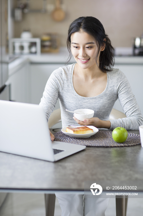 年轻女子一边吃早餐一边使用笔记本电脑