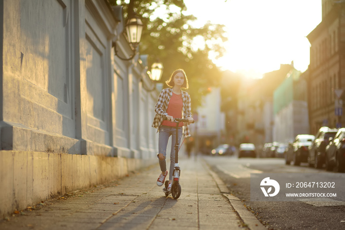 Adorable girl riding her scooter in a city park on sunny autumn evening. Pretty preteen child riding