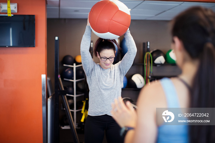 Rear view of female athlete looking at friend exercising with medicine ball while standing in gym