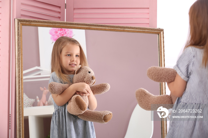 Cute little girl with teddy bear looking in mirror at home