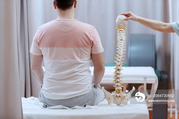 Male patient sitting on the hospital bed with backs turned. Next to him is nurse holding spine model