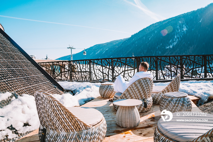 Young man in a robe chilling on a open terrace in the SPA with an amazing mountains view during wint