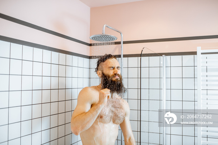 Bearded man feeling shocked while taking a shower with cold water in the bathroom at home