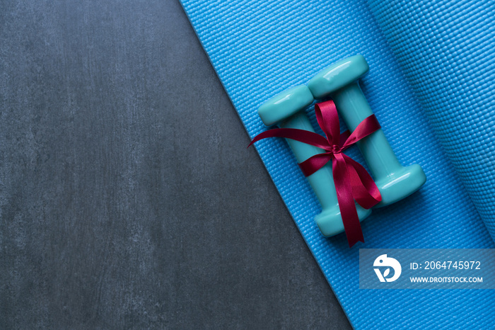 两个绿色哑铃，蓝色瑜伽垫背景上有红色礼物蝴蝶结，运动和健康的概念