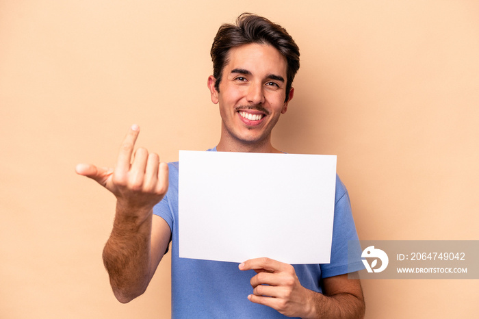 Young caucasian man holding a placard isolated on beige background pointing with finger at you as if