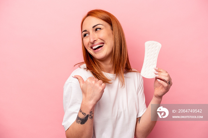 Young caucasian woman holding sanitary napkin isolated on pink background points with thumb finger a