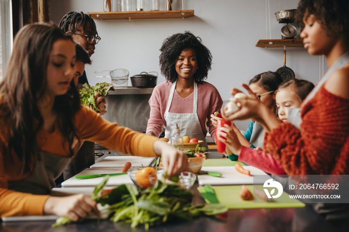 Smiling female chef looking at students holding vegetables on table while standing against wall in c