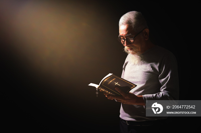 senior man standing in dark background holding and reading holy bible