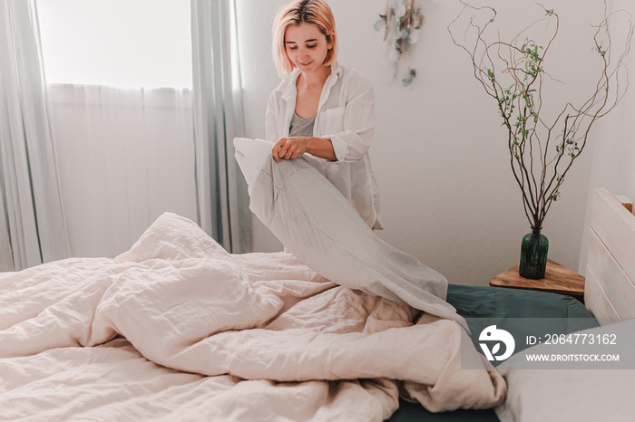 Young pretty woman is making a bed. Fresh bedding linen. Put on pillow cover. Light and bright room.