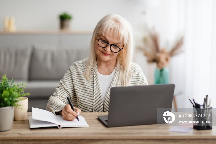 Mature lady writing in notebook and using laptop at home