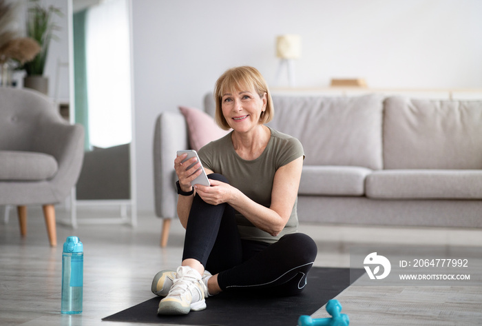 Active senior woman taking break, checking her smartphone on yoga mat after home workout