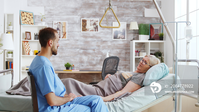 Male assistant taking a seat near hospital bed of ill lady lying in nursing home