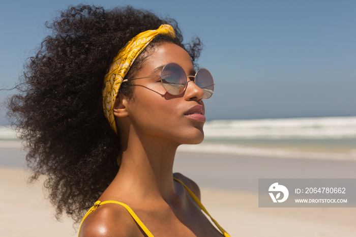 Young African American woman in yellow bikini and sunglasses standing on the beach