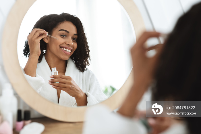 Smiling black woman applying moisturising serum to face and looking to mirror