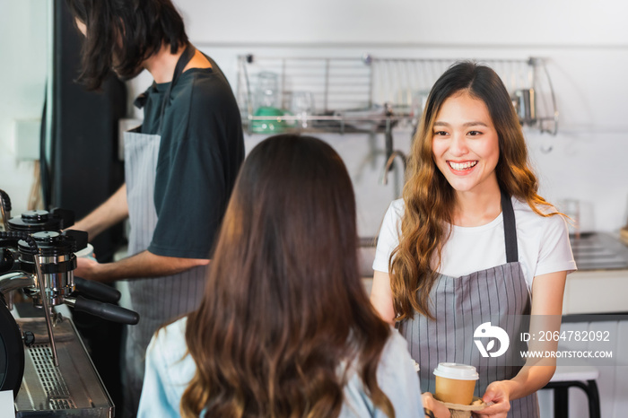 年轻漂亮的亚洲女咖啡师系着围裙，手里拿着热咖啡杯在酒吧为顾客服务