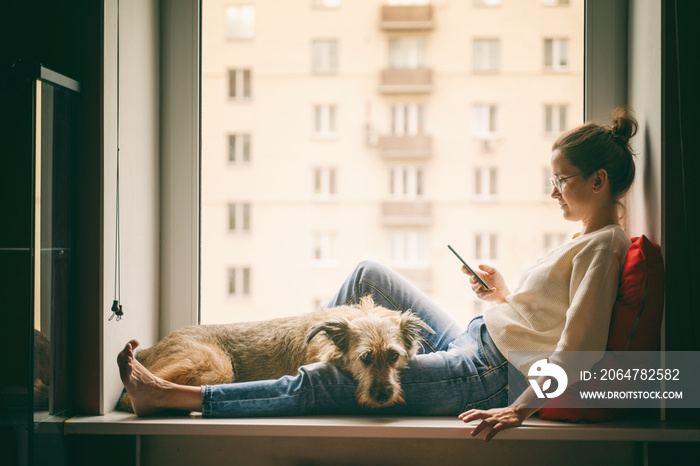 A young woman with her brown dog is sitting on a windowsill overlooking the city, looking at the sma