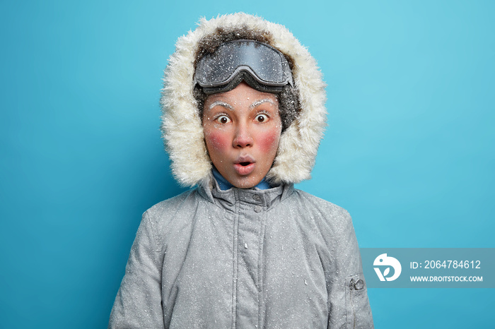 Shocked female skier with red frozen face stares at camera dressed in grey jacket with hood and snow