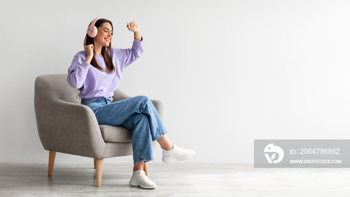 Cheery young lady in headphones listeing to music and dancing while sitting in armchair against whit