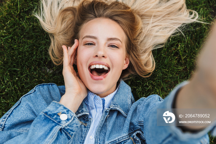 Photo of blonde kind woman taking selfie photo with hand on her face while lying green grass in park