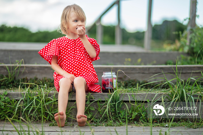 Little girl with glass jar of fresh berries. Blonde hair and blue eyes kid in polka dot red dress. S